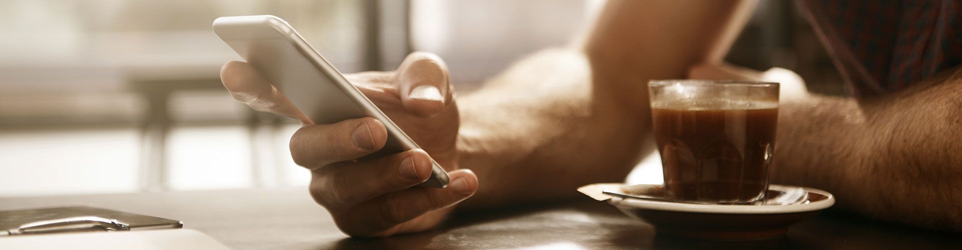 Foto: Hand die auf dem Tisch liegt und ein Handy hält. Im Vordergrund eine Kaffetasse-